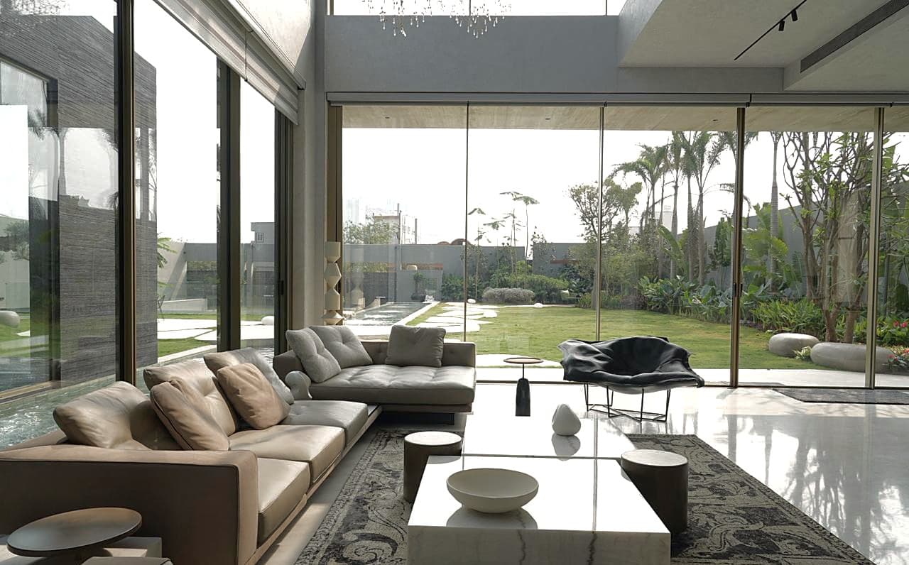 Living room opening to the garden: taupe and grey L-shaped sofas, marble block tables with a stone bowl, sculptural lounge chair against palms.