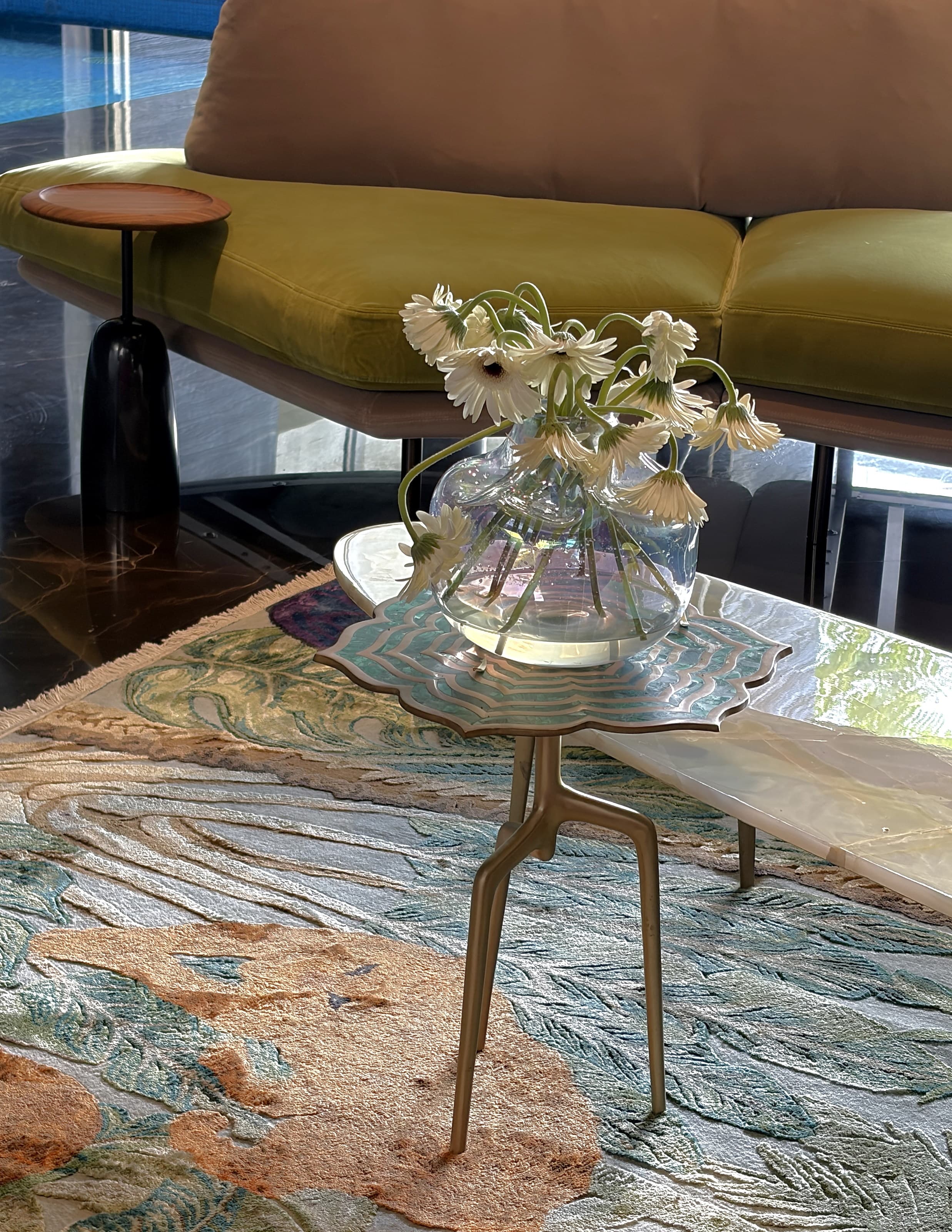 Gerberas in an iridescent glass vase on a scalloped side table, embroidered rug below.