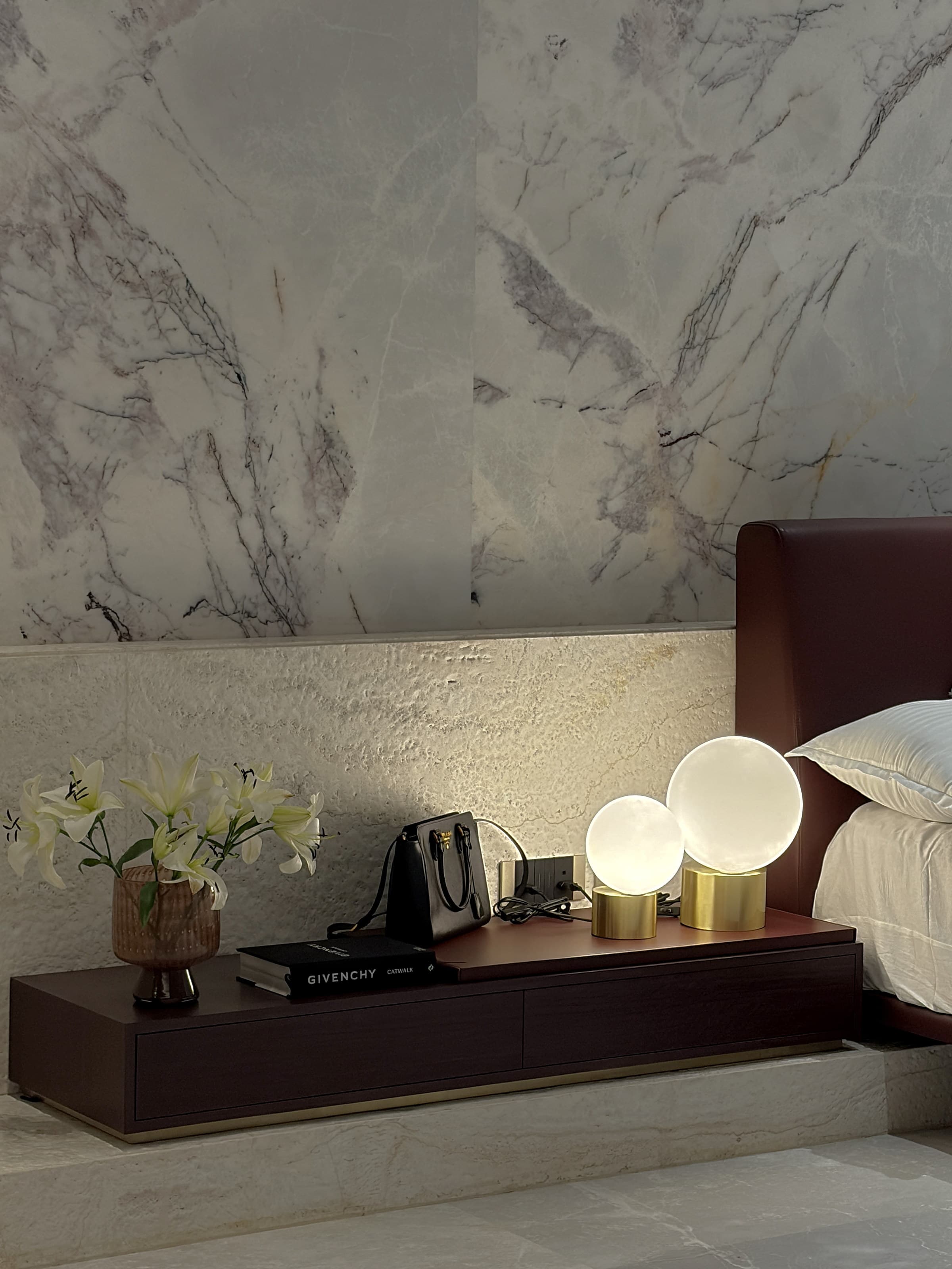 Bedside still life: lilies, stacked monographs and two brass globe lamps on a burgundy console.