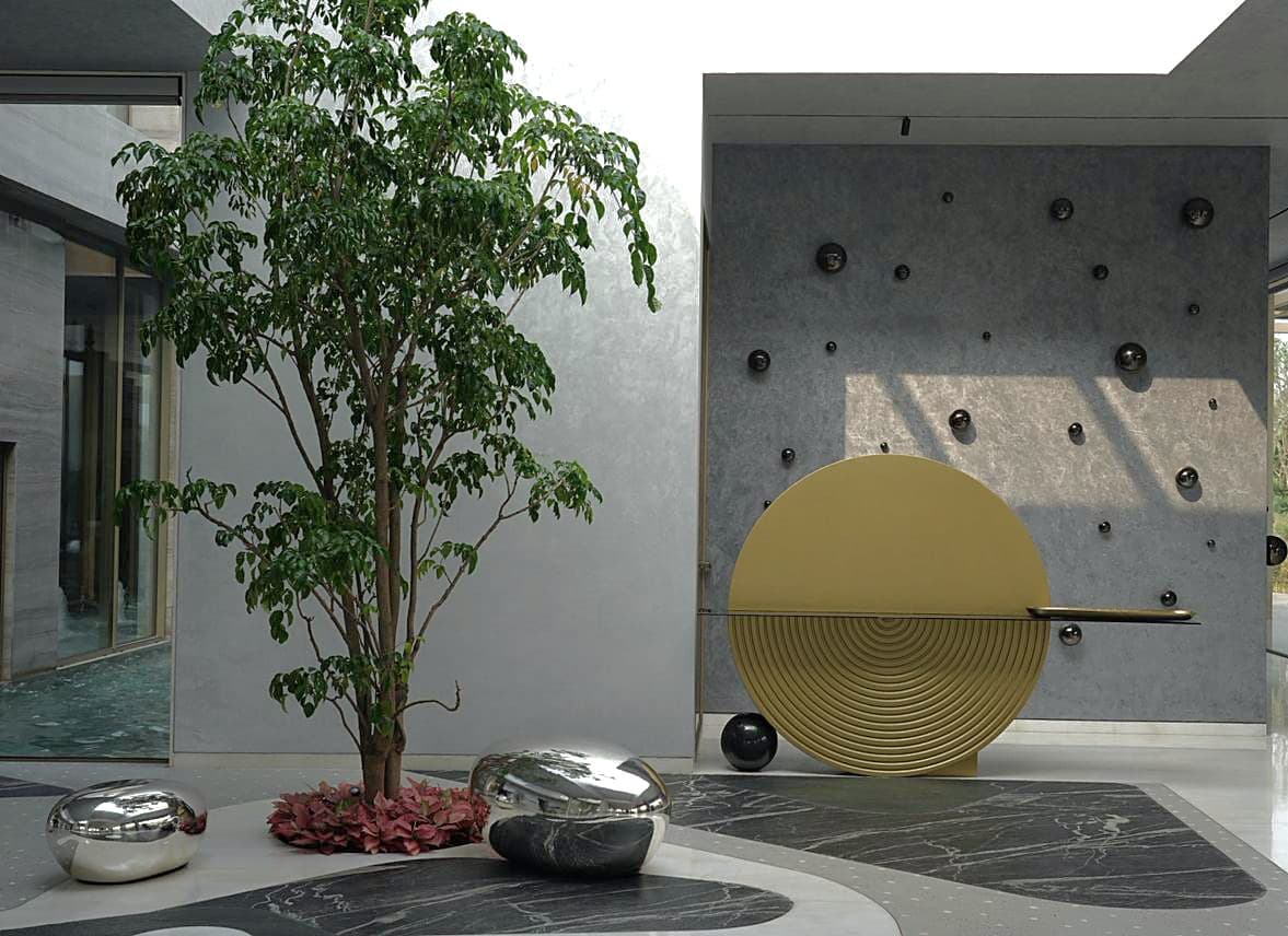 Courtyard: gold ridged disc console against a studded wall, mirror pebbles and the tree in dappled sun.