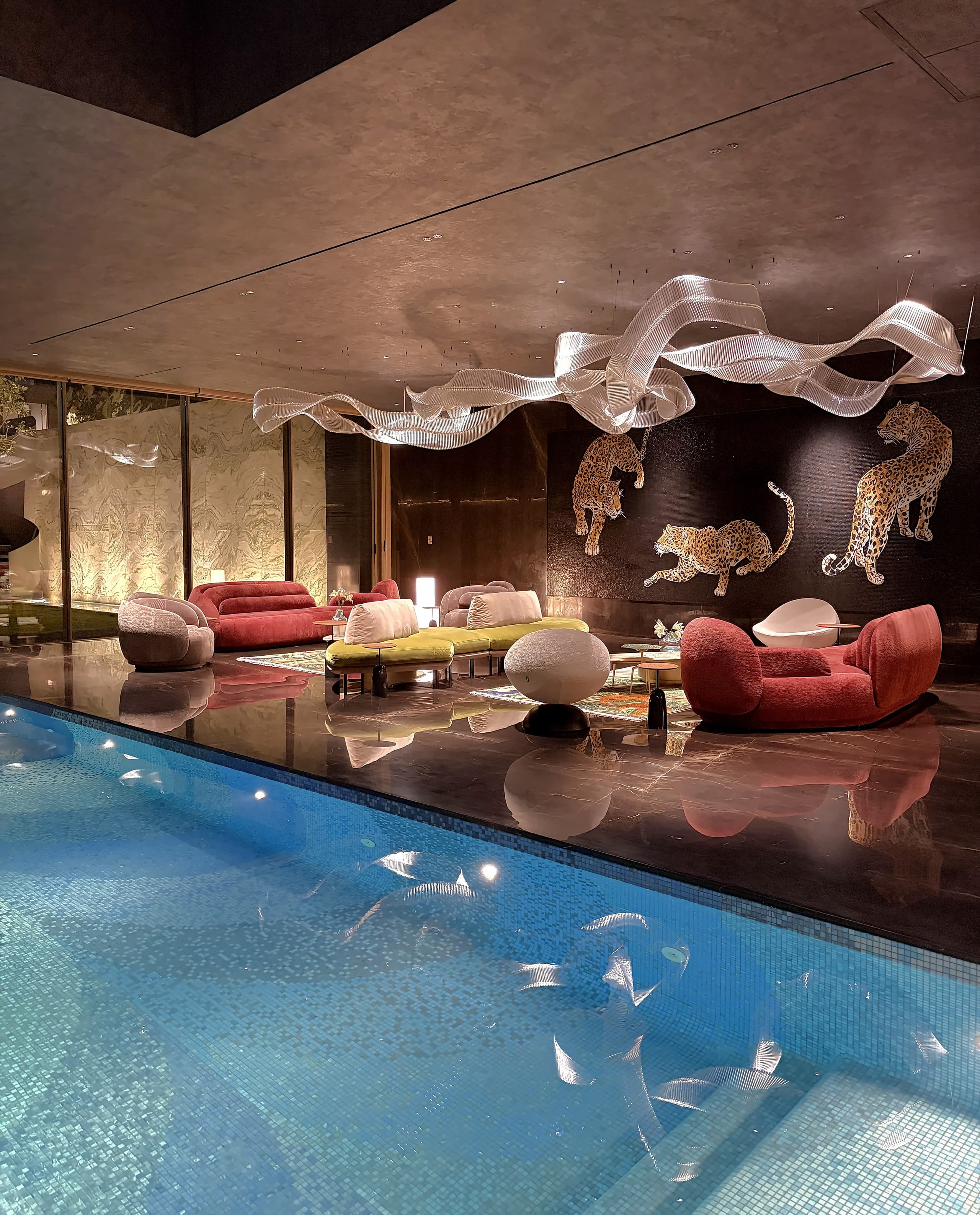 The same lounge at night: leopard wall, glass ribbon chandelier, rose and chartreuse seating mirrored in the pool.