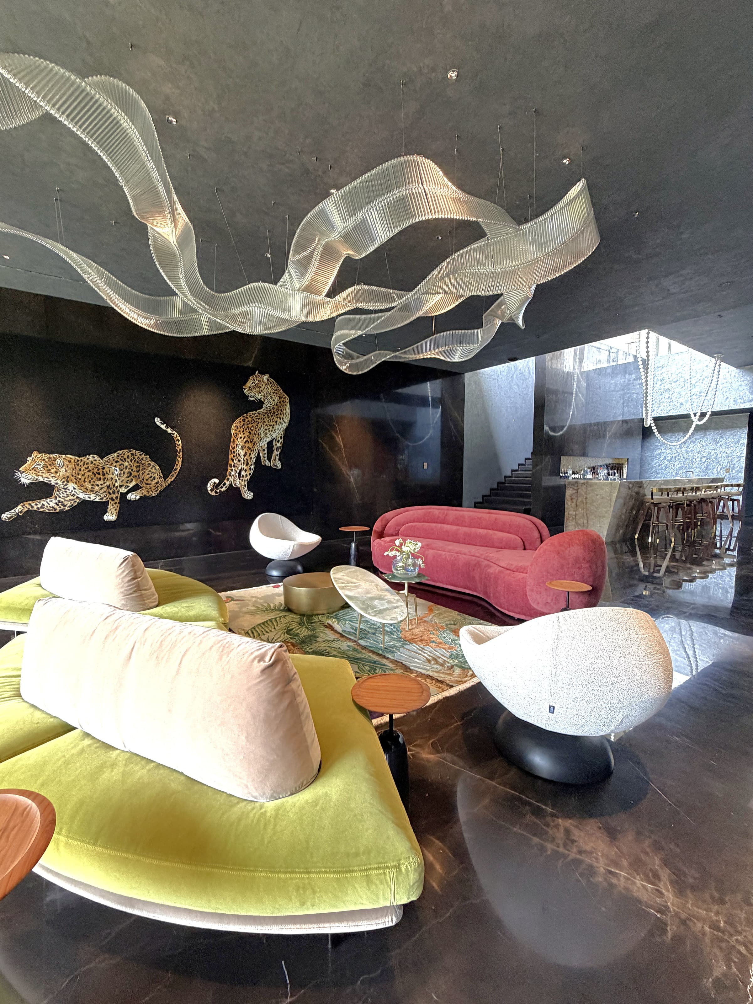Pool lounge in daylight: chartreuse and rose sofas, white sculptural chairs, leopard wall and the glass ribbon chandelier overhead.
