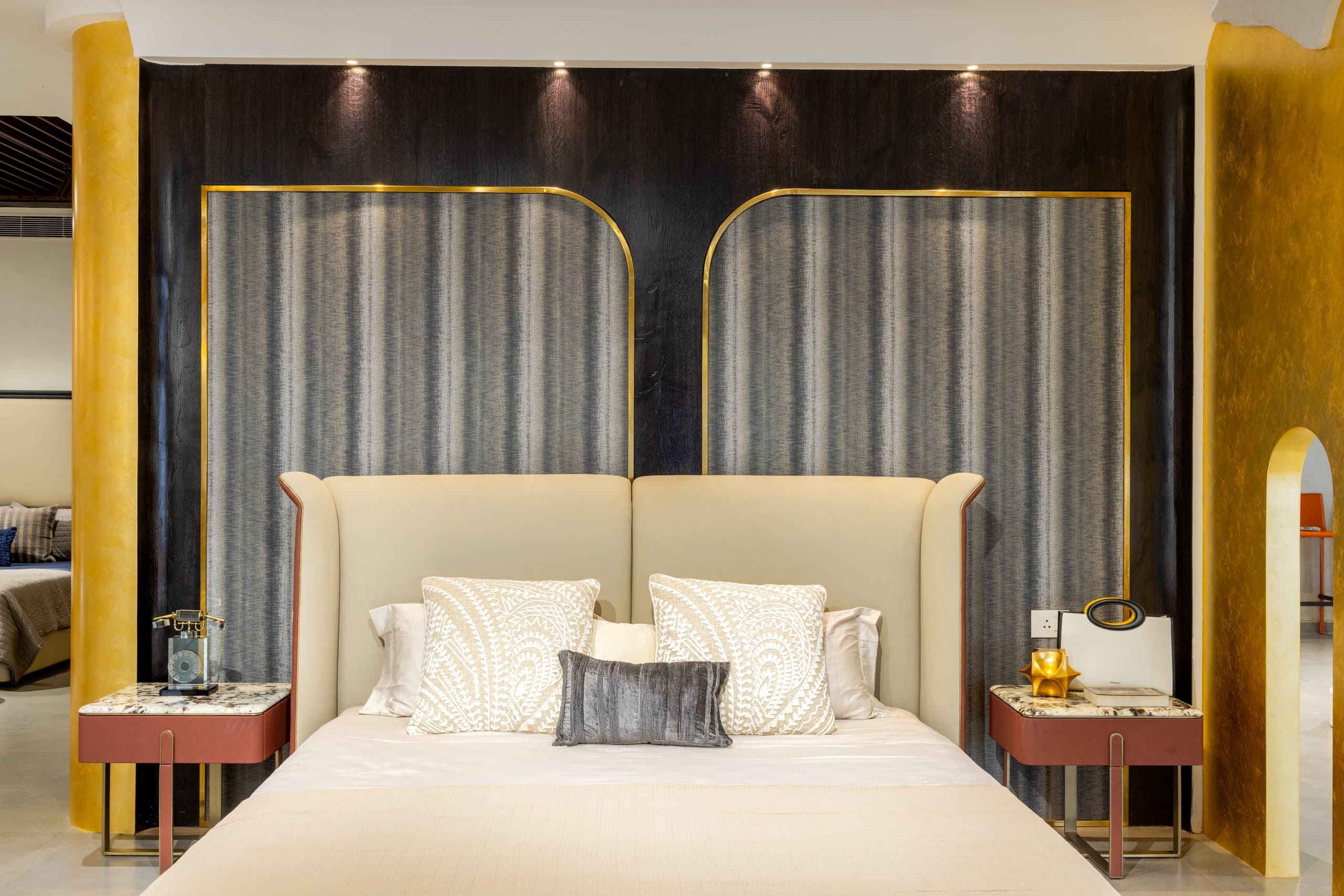 Bedside detail: cream winged headboard with oxblood piping against brass-trimmed fabric wall panels, embroidered cushions, and a burgundy marble-topped nightstand holding a vintage telephone.