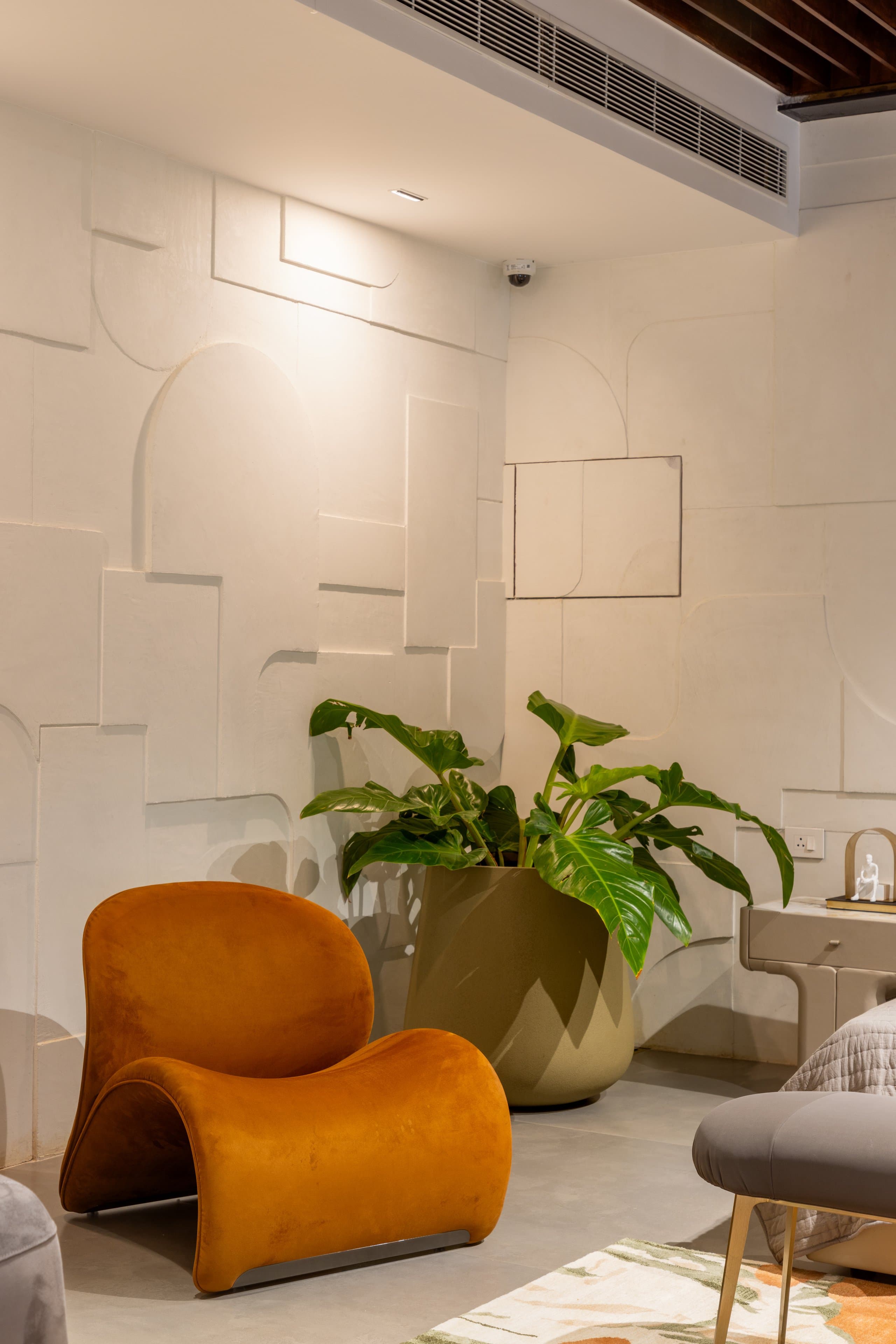 Mustard velvet sculptural lounge chair against a white geometric relief-plaster wall, with a large plant in an olive-green planter.