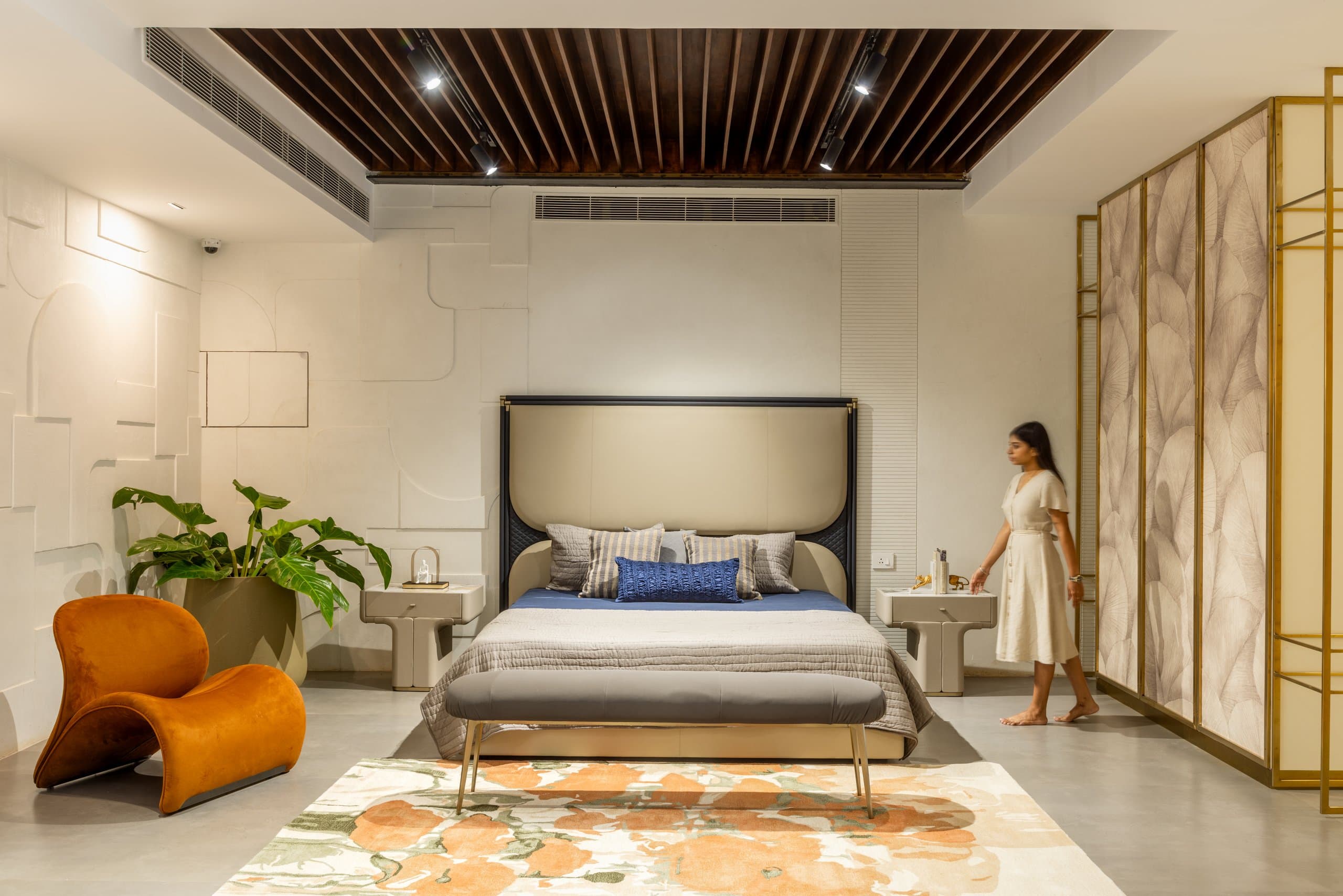 Bedroom vignette beneath a timber-slat ceiling: cream and navy bed with a blue lumbar cushion, grey bench, floral rug in orange and green, brass-framed floral screen, and an orange sculptural chair nearby.