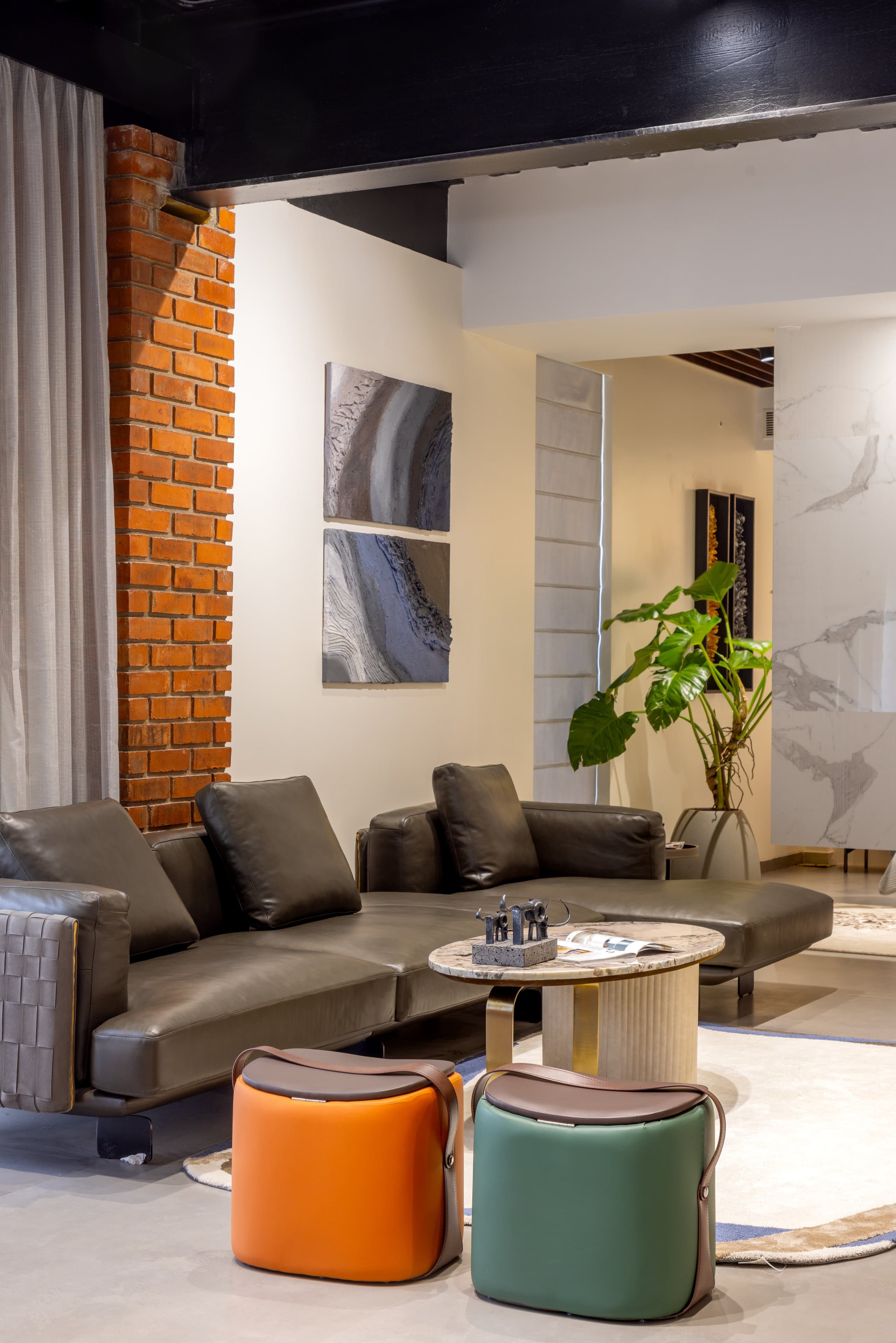 Green-grey leather sofa with woven arms beneath two stacked abstract canvases, beside a round marble-and-brass table and a pair of leather-strapped poufs in orange and green.