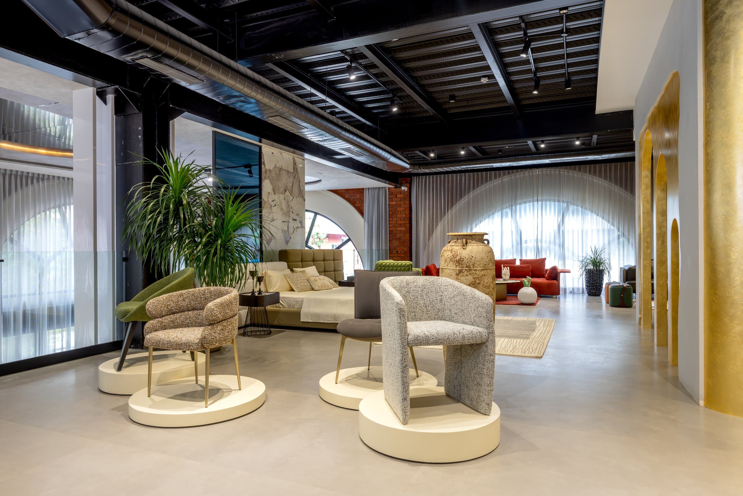 Four accent chairs displayed one per cream plinth — green bouclé, speckled beige, grey, and grey-black — with plants and showroom room-settings visible beyond.