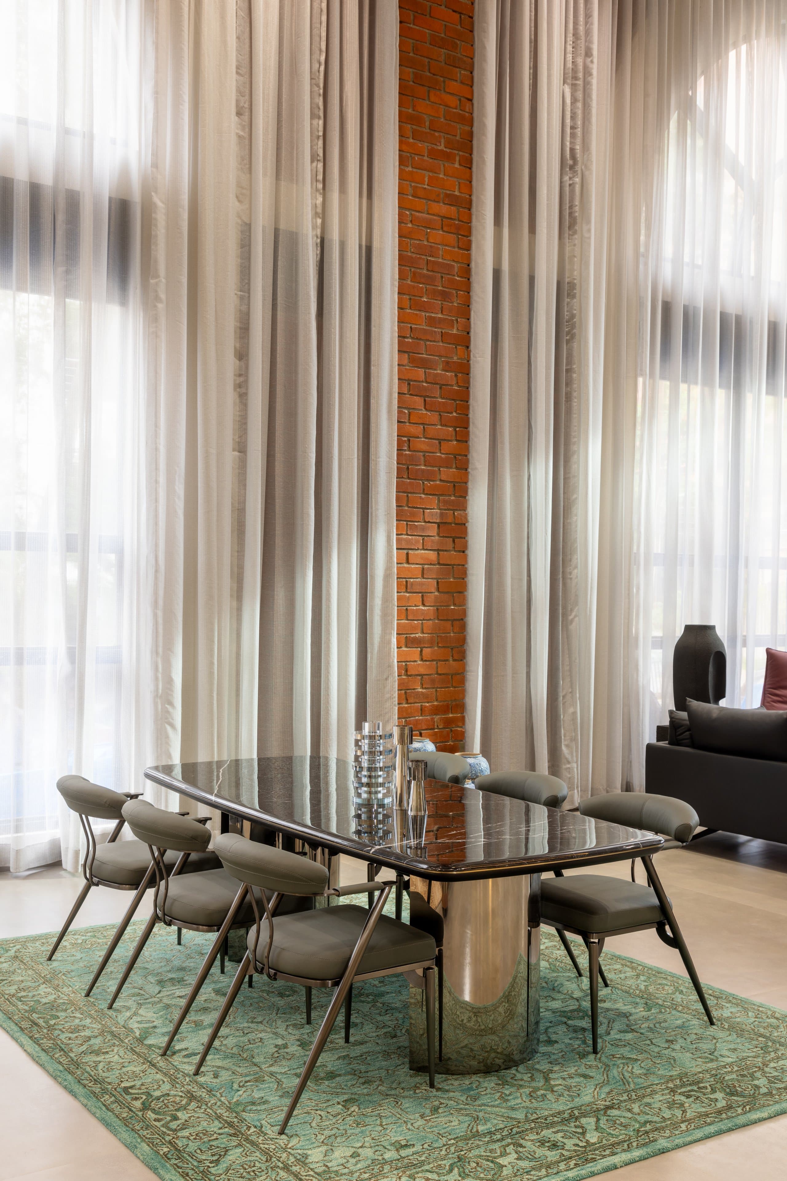 Oval black marble dining table on a polished steel base with six grey leather chairs, standing on a teal overdyed rug in front of a brick pier and full-height sheer curtains.
