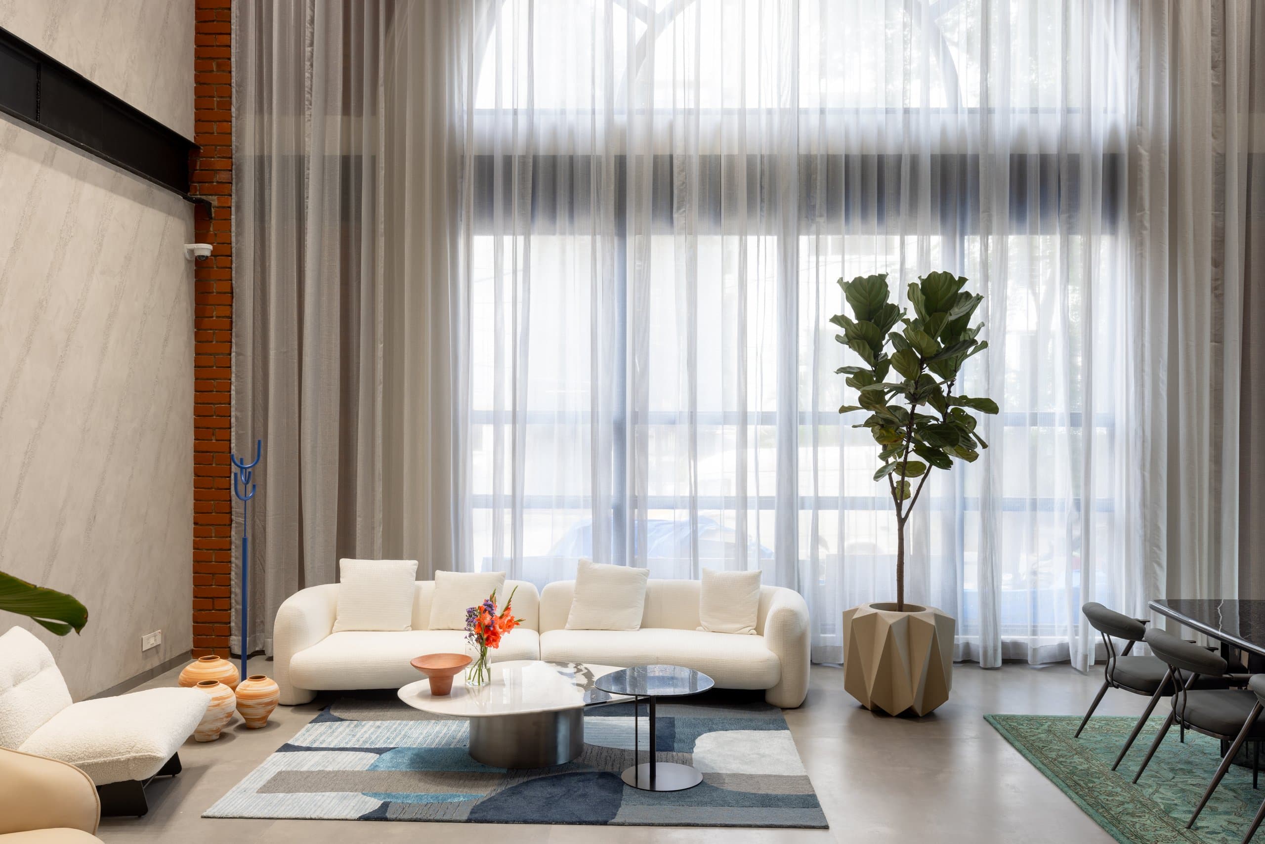 Showroom lounge: two curved white sofas around stone and smoked-glass tables on a blue patchwork rug, a cobalt-blue sculptural candelabra, a fiddle-leaf fig in a faceted planter and tall sheer curtains behind.