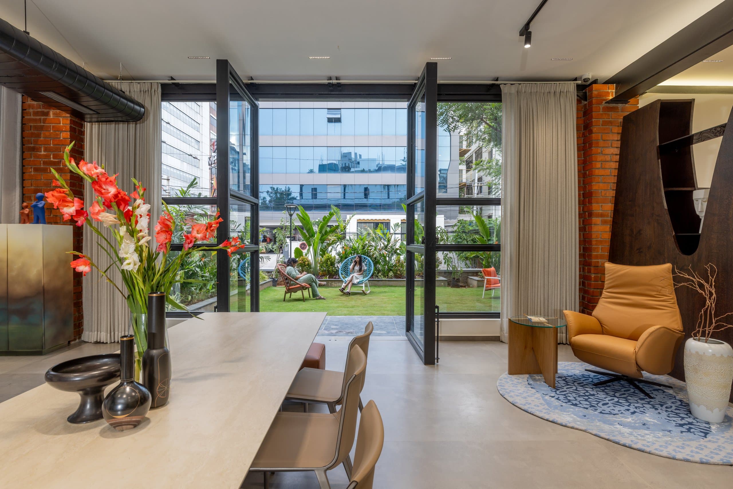 Travertine dining table set with three black vessels and a vase of red gladioli, tan leather chairs, and steel-framed glass doors open to a garden with two wire chairs on the lawn.