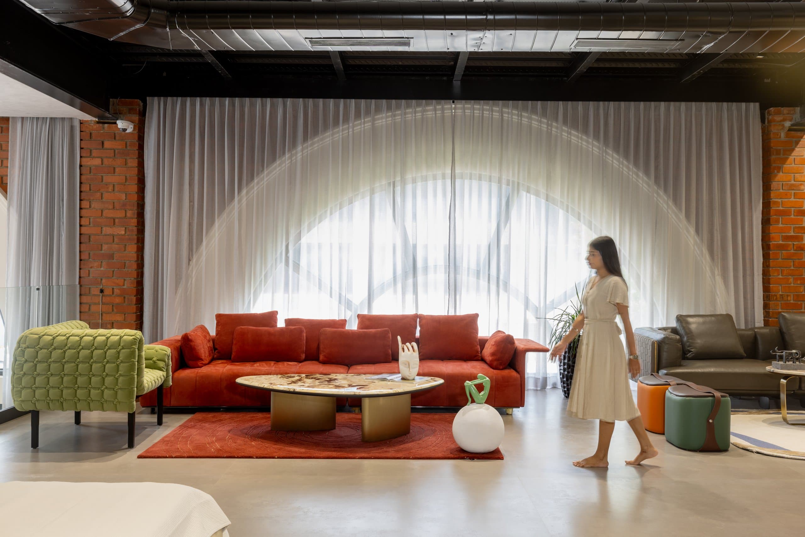 A red territory in the showroom: red velvet sofa with red cushions on a red rug beneath an arched sheer-curtained window, faced by a green quilted armchair, with a stone-topped coffee table and white sphere sculpture.