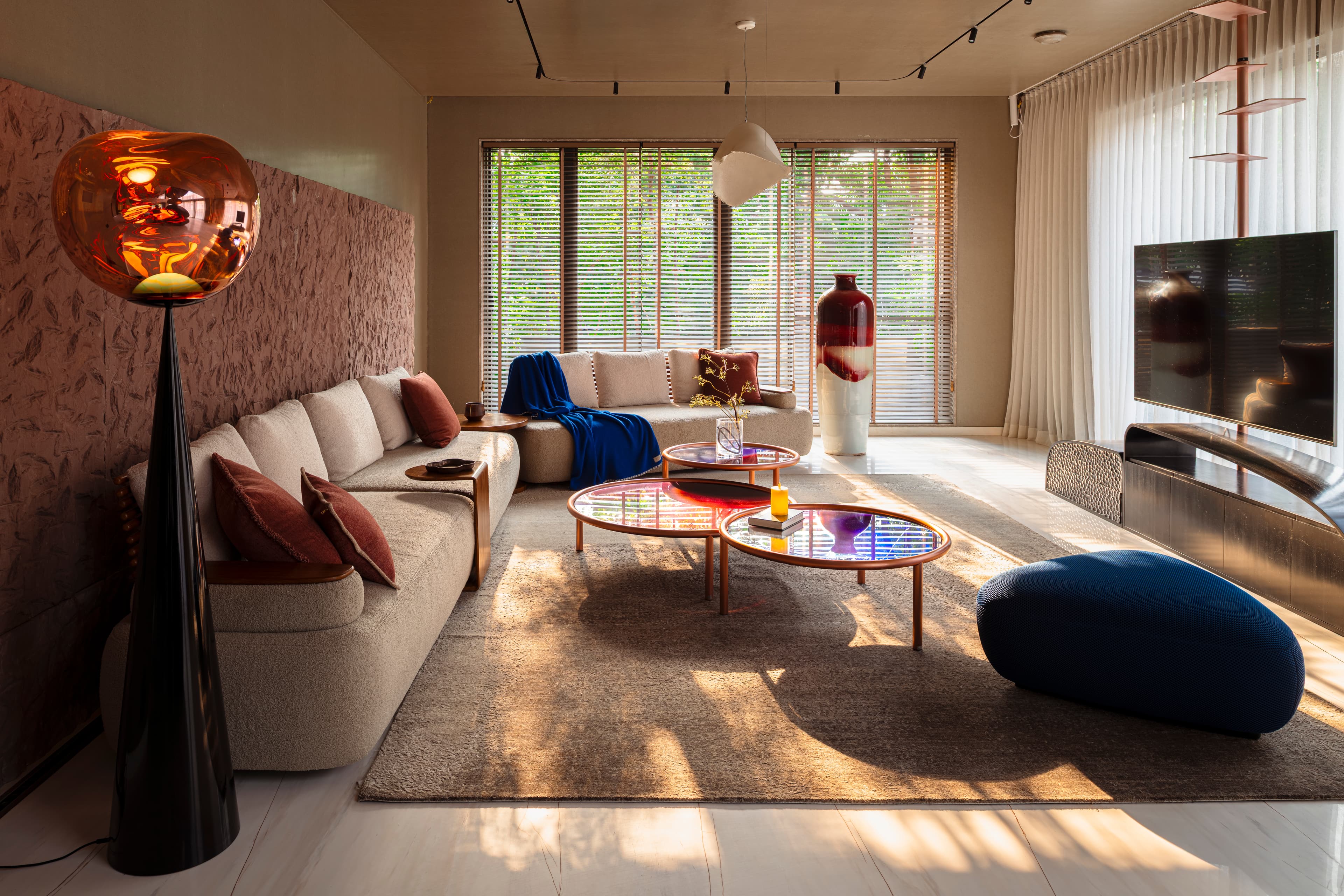 Family lounge in evening light: ivory sofa with a cobalt-blue throw and rust cushions, a copper globe floor lamp, round glass-topped nesting tables and a blue pebble-shaped pouf on a large rug.